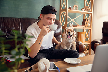 Image showing Young handsome man sitting on sofa at home with his cute dog. Cozy office workplace, remote work, online, e-learning concept.