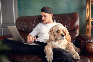 Image showing Young handsome man sitting and working at home with his cute dog. Cozy office workplace, remote work, online learning concept.