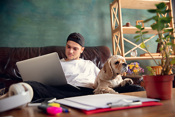 Image showing Young handsome man sitting and working at home with his cute dog. Cozy office workplace, remote work, online learning concept.