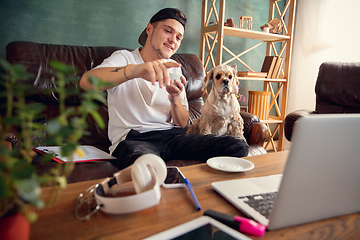 Image showing Young man sitting on sofa at home with cute dog and chatting with his friends online.