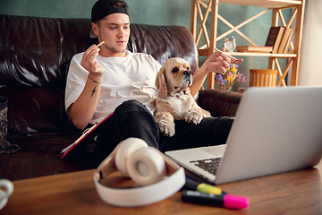 Image showing Young man sitting on sofa at home with cute dog and chatting with his friends online.