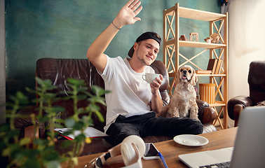 Image showing Young man sitting on sofa at home with cute dog and chatting with his friends online.