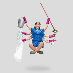 Image showing Young woman housemaid in headphones and blue uniform multitask like shiva isolated on gray background.