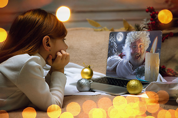 Image showing Happy caucasian little girl during video call or messaging with Santa using laptop and home devices, celebration or ad flyer with copyspace