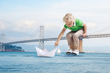 Image showing See the world by children\'s eyes - little cute curly boy playing with paper ship sitting on bridge