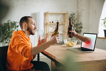 Image showing Young man drinking beer during meeting friends on virtual video call. Distance online meeting, chat together on laptop at home.