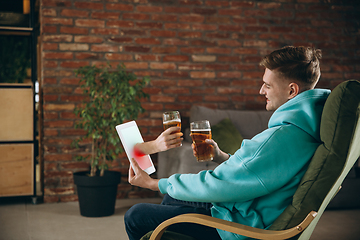 Image showing Young man drinking beer during meeting friends on virtual video call. Distance online meeting, chat together on tablet at home.