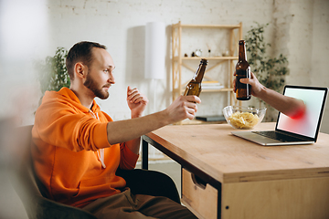 Image showing Young man drinking beer during meeting friends on virtual video call. Distance online meeting, chat together on laptop at home.