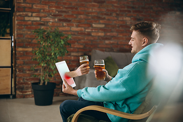 Image showing Young man drinking beer during meeting friends on virtual video call. Distance online meeting, chat together on tablet at home.