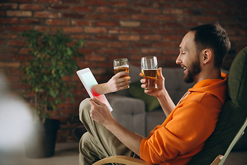 Image showing Young man drinking beer during meeting friends on virtual video call. Distance online meeting, chat together on tablet at home.
