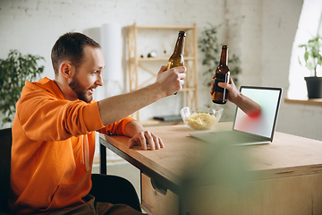 Image showing Young man drinking beer during meeting friends on virtual video call. Distance online meeting, chat together on laptop at home.