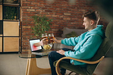 Image showing Young man drinking beer during meeting friends on virtual video call. Distance online meeting, chat together on laptop at home.