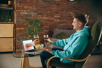 Image showing Young man drinking beer during meeting friends on virtual video call. Distance online meeting, chat together on laptop at home.