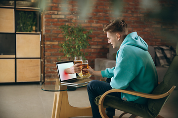 Image showing Young man drinking beer during meeting friends on virtual video call. Distance online meeting, chat together on laptop at home.