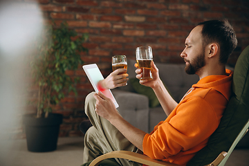 Image showing Young man drinking beer during meeting friends on virtual video call. Distance online meeting, chat together on tablet at home.