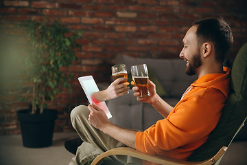 Image showing Young man drinking beer during meeting friends on virtual video call. Distance online meeting, chat together on tablet at home.