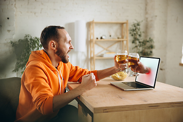 Image showing Young man drinking beer during meeting friends on virtual video call. Distance online meeting, chat together on laptop at home.