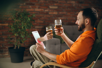 Image showing Young man drinking beer during meeting friends on virtual video call. Distance online meeting, chat together on tablet at home.