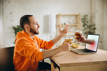 Image showing Young man drinking beer during meeting friends on virtual video call. Distance online meeting, chat together on laptop at home.