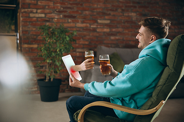 Image showing Young man drinking beer during meeting friends on virtual video call. Distance online meeting, chat together on tablet at home.