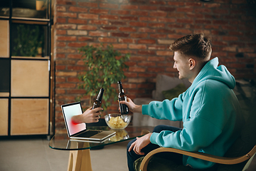 Image showing Young man drinking beer during meeting friends on virtual video call. Distance online meeting, chat together on laptop at home.