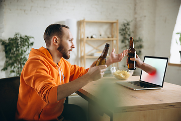 Image showing Young man drinking beer during meeting friends on virtual video call. Distance online meeting, chat together on laptop at home.