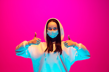 Image showing Caucasian beautiful woman\'s portrait isolated on pink studio background in multicolored neon light. Wearing face mask.