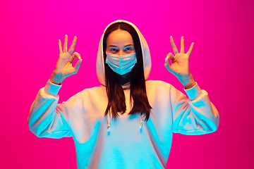 Image showing Caucasian beautiful woman\'s portrait isolated on pink studio background in multicolored neon light. Wearing face mask.