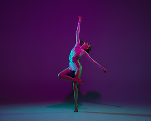 Image showing Young female athlete, rhythmic gymnastics artist on purple background with neon light. Beautiful girl practicing with equipment. Grace in performance.