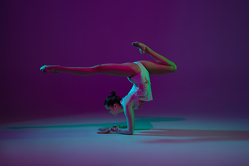 Image showing Young female athlete, rhythmic gymnastics artist on purple background with neon light. Beautiful girl practicing with equipment. Grace in performance.