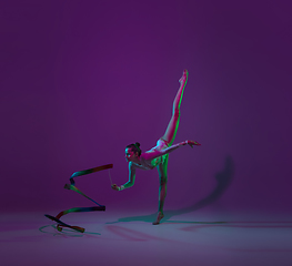 Image showing Young female athlete, rhythmic gymnastics artist on purple background with neon light. Beautiful girl practicing with equipment. Grace in performance.