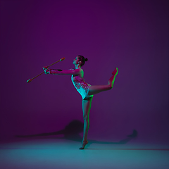 Image showing Young female athlete, rhythmic gymnastics artist on purple background with neon light. Beautiful girl practicing with equipment. Grace in performance.