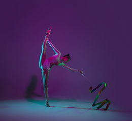 Image showing Young female athlete, rhythmic gymnastics artist on purple background with neon light. Beautiful girl practicing with equipment. Grace in performance.