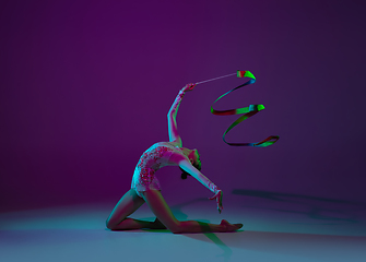 Image showing Young female athlete, rhythmic gymnastics artist on purple background with neon light. Beautiful girl practicing with equipment. Grace in performance.