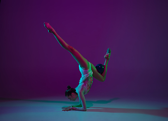 Image showing Young female athlete, rhythmic gymnastics artist on purple background with neon light. Beautiful girl practicing with equipment. Grace in performance.