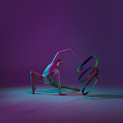 Image showing Young female athlete, rhythmic gymnastics artist on purple background with neon light. Beautiful girl practicing with equipment. Grace in performance.