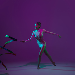 Image showing Young female athlete, rhythmic gymnastics artist on purple background with neon light. Beautiful girl practicing with equipment. Grace in performance.