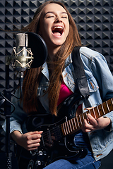 Image showing Girl musician in recording studio playing electric guitar and singing in mic