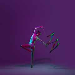 Image showing Young female athlete, rhythmic gymnastics artist on purple background with neon light. Beautiful girl practicing with equipment. Grace in performance.