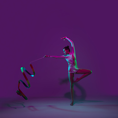 Image showing Young female athlete, rhythmic gymnastics artist on purple background with neon light. Beautiful girl practicing with equipment. Grace in performance.