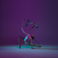 Image showing Young female athlete, rhythmic gymnastics artist on purple background with neon light. Beautiful girl practicing with equipment. Grace in performance.