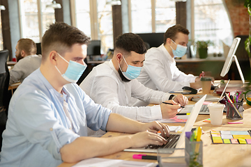 Image showing Young caucasian colleagues working together in a office using modern devices and gadgets during quarantine. Wearing protective face masks