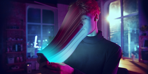 Image showing Cinematic portrait of stylish man in neon lighted interior using a smartphone. The face is smeared, sucked into the phone. The concept of social network dependency