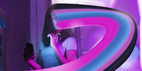 Image showing Cinematic portrait of stylish woman in neon lighted interior using a smartphone. The face is smeared, sucked into the phone. The concept of social network dependency