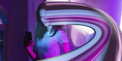 Image showing Cinematic portrait of stylish woman in neon lighted interior using a smartphone. The face is smeared, sucked into the phone. The concept of social network dependency