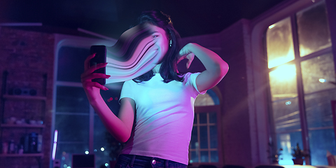 Image showing Cinematic portrait of stylish woman in neon lighted interior using a smartphone. The face is smeared, sucked into the phone. The concept of social network dependency