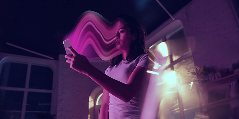 Image showing Cinematic portrait of stylish woman in neon lighted interior using a smartphone. The face is smeared, sucked into the phone. The concept of social network dependency