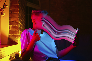 Image showing Cinematic portrait of stylish woman in neon lighted interior using a smartphone. The face is smeared, sucked into the screen. The concept of social network dependency