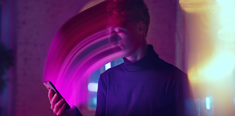 Image showing Cinematic portrait of stylish man in neon lighted interior using a smartphone. The face is smeared, sucked into the phone. The concept of social network dependency