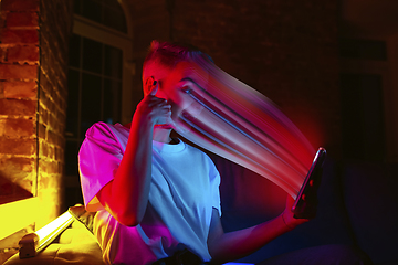 Image showing Cinematic portrait of stylish woman in neon lighted interior using a smartphone. The face is smeared, sucked into the phone. The concept of social network dependency